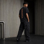 A man stands in a concrete corridor wearing a black t-shirt with a vertical orange graphic on the back, black cargo pants, and black sneakers.