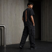 A man stands in a concrete corridor wearing a black t-shirt with a vertical orange graphic on the back, black cargo pants, and black sneakers.