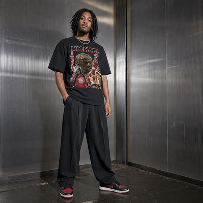 A man wearing a black graphic t-shirt with a Michael Jordan design, black sweatpants, and red and black sneakers, standing against a metallic gray wall.
