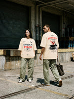 A man and a woman wearing matching beige graphic t-shirts and olive green pants walking in an industrial outdoor setting with closed metal garage doors and concrete ground.