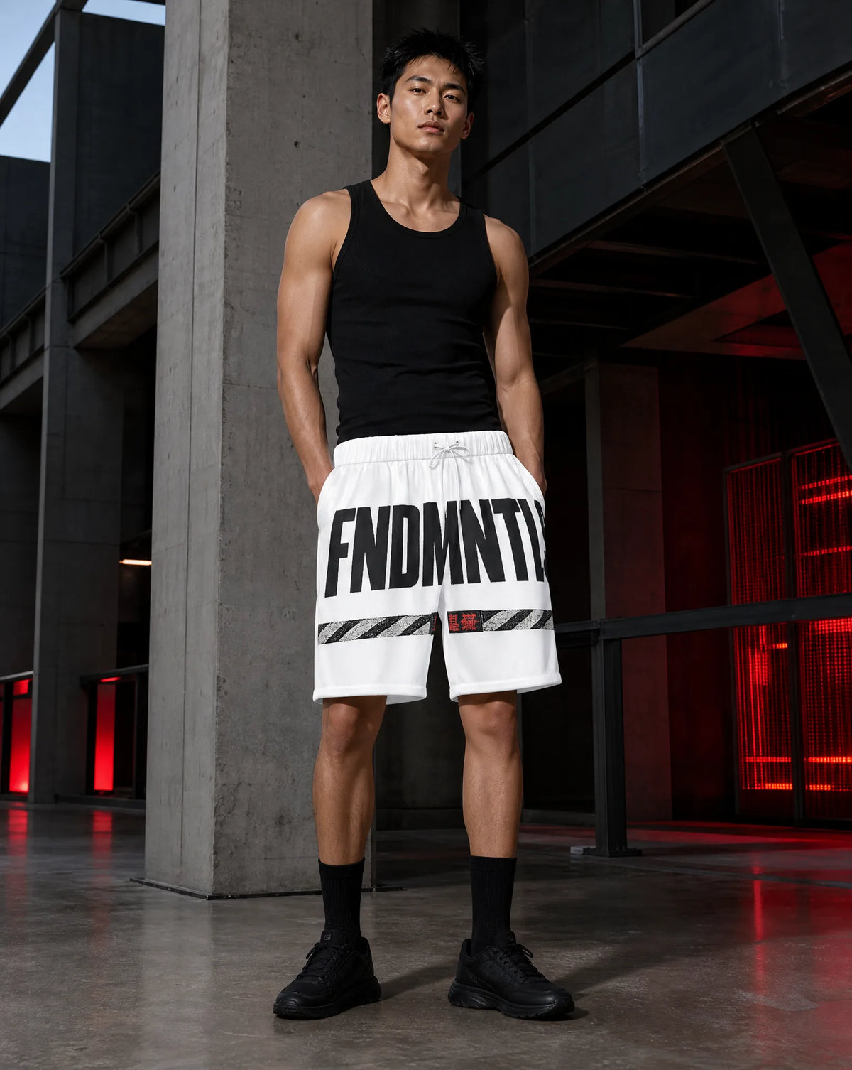 A man stands in a modern industrial setting wearing white athletic shorts with bold black text reading 'FNDMNTLS', a black tank top, black socks, and black sneakers.