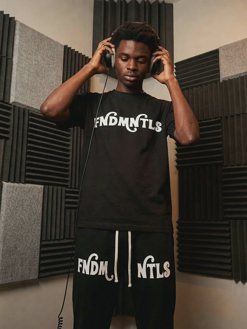 Mr Rogers FNDMNTLS Flared Sweatpants styled in a music studio with matching black tee, showcasing bold white logo and relaxed fit.