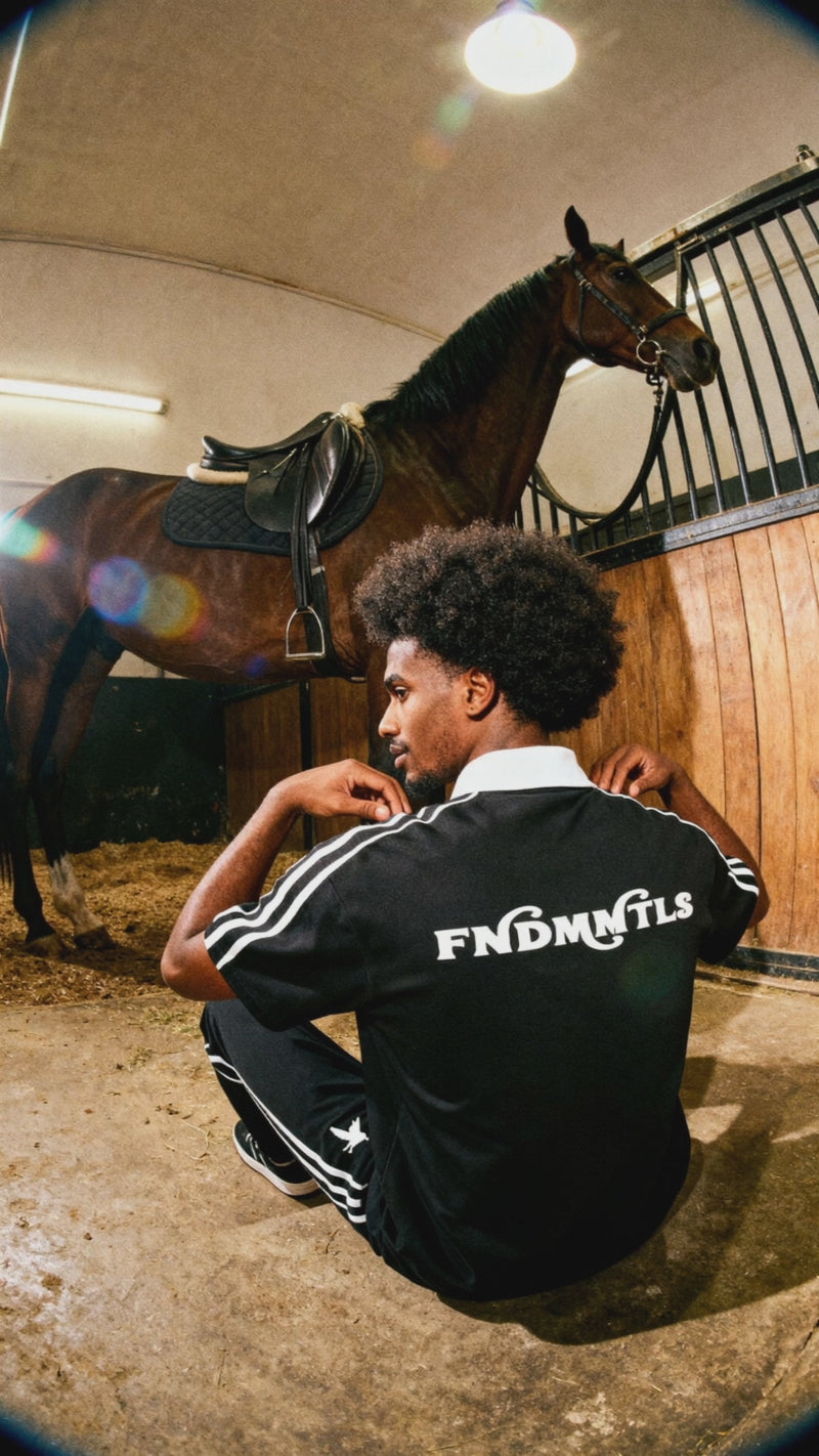 Short-Sleeve Polo Shirt - Contrast Collar | Cotton Blend in black with bold FNDMNTLS logo, worn in a rustic stable setting with a saddled horse.